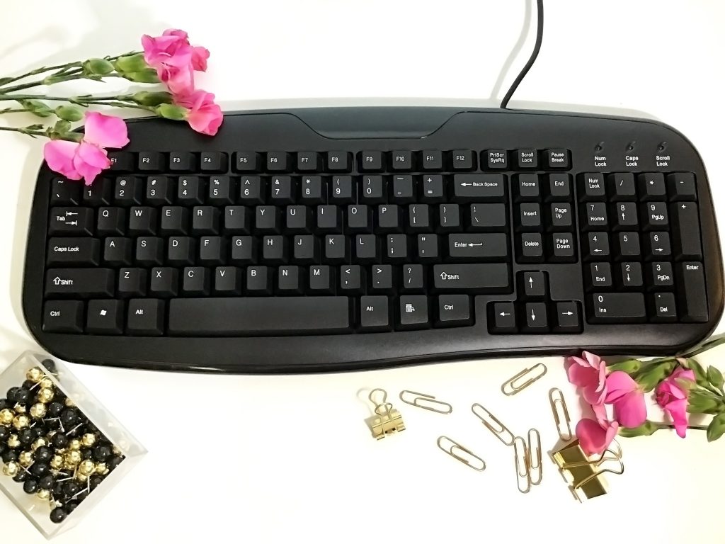 Styled Stock Photo of a black computer keyboard with pink flowers, gold paperclips, and black and gold thumb tacks on a white background. Become a Member to the Best Stock Photography Site SwankyStock